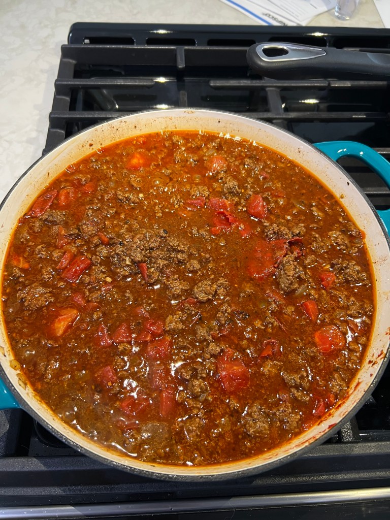 A large pot of simmering chili with ground beef and diced tomatoes on a stovetop.