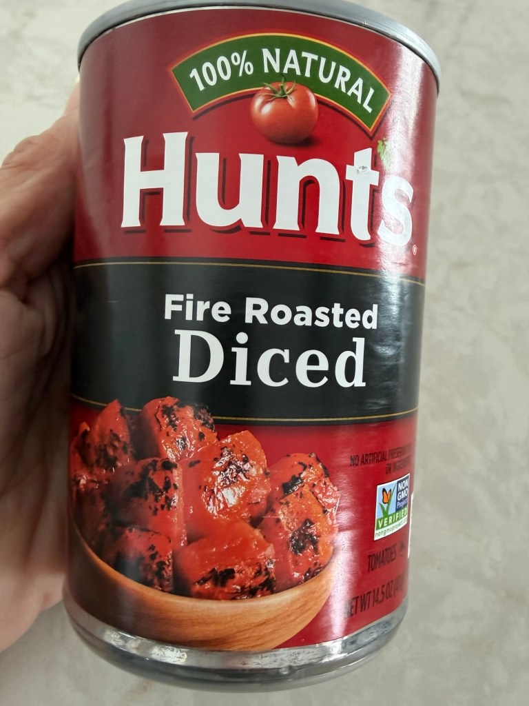 A hand holding a can of Hunt's Fire Roasted Diced Tomatoes, featuring a red label stating '100% Natural' and showcasing diced tomatoes in a bowl on the can.
